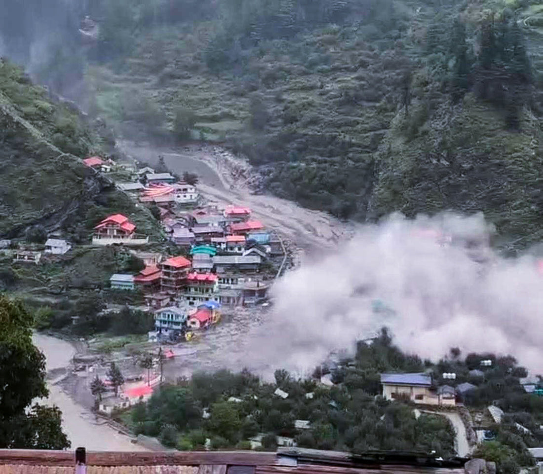 Muddy floodwaters surging through the hillside village of Dharali after a sudden cloudburst, sweeping debris past stone houses under dark storm clouds