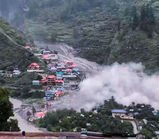 Muddy floodwaters surging through the hillside village of Dharali after a sudden cloudburst, sweeping debris past stone houses under dark storm clouds