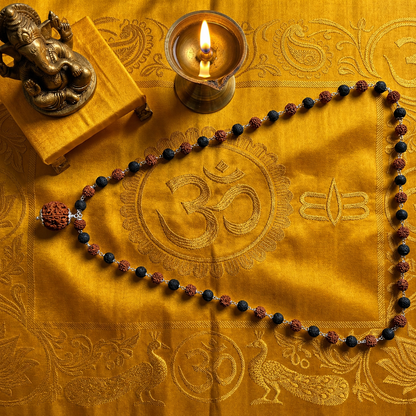 Karungali Rudraksha mala Silver Chain on decorative table setting with a rosary, candle, and small statue on a yellow embroidered cloth.
